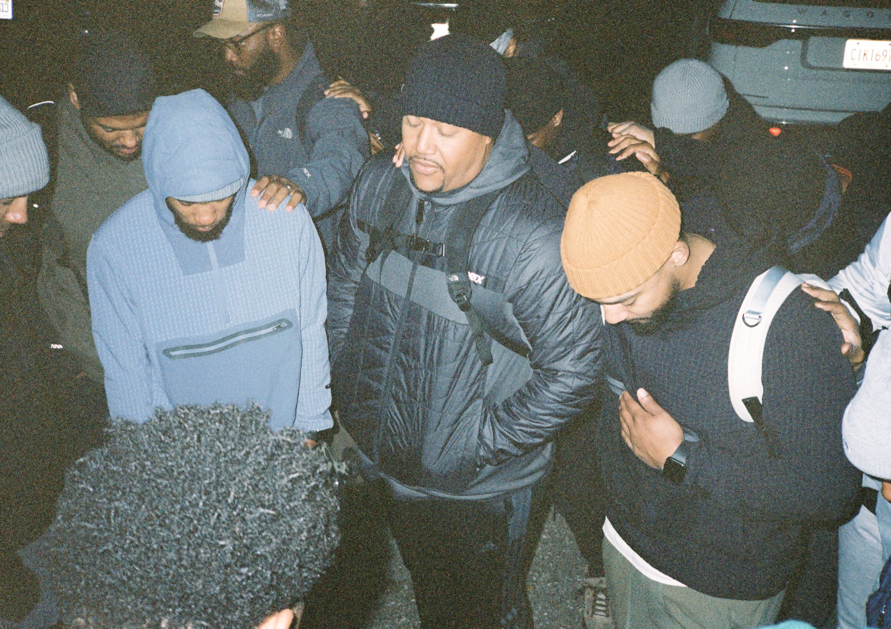 Men standing in a circle at night, heads bowed, hands resting on each other's shoulders.